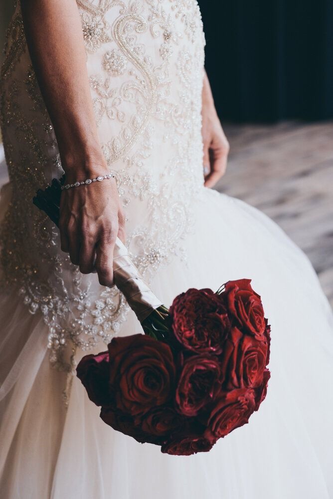 Bride in a beaded wedding gown holds a bouquet of red roses.
