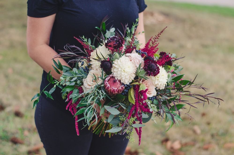 Woman holding a vibrant bouquet with white, burgundy, and green flowers outdoors.