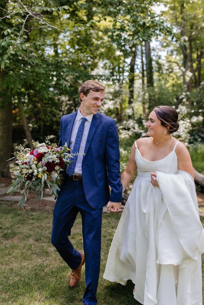 Newlyweds walk hand-in-hand in a wooded area; the groom in a blue suit, the bride in a white dress, smiling at each other.