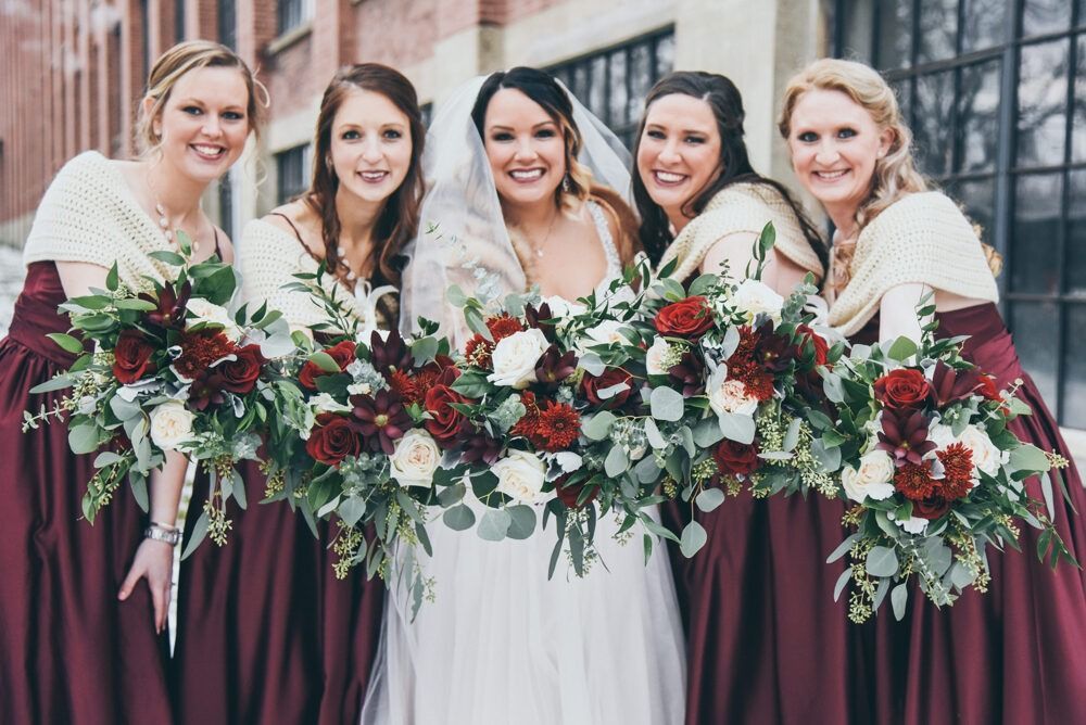 Bride and bridesmaids holding bouquets, wearing burgundy dresses and shawls, smiling outdoors.