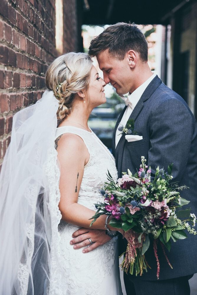 Bride and groom embrace by a brick wall, she in a white dress and veil, he in a suit, holding bouquet.