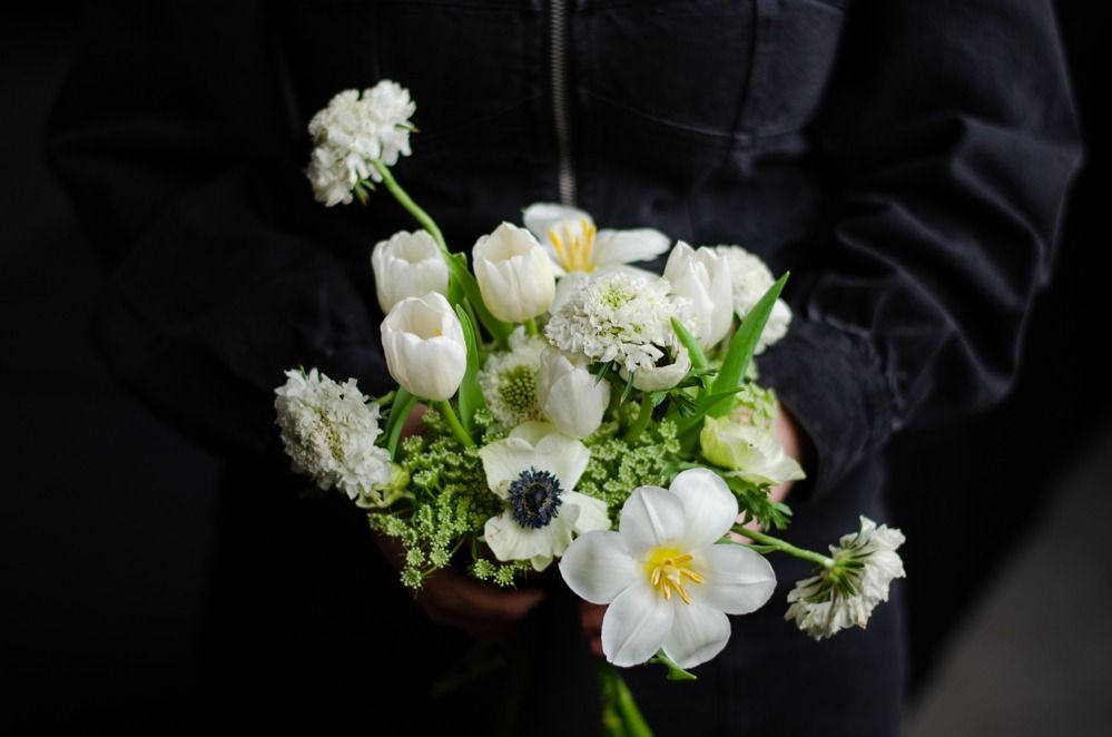 Person holding a bouquet of white flowers, including tulips and anemones, against a dark background.