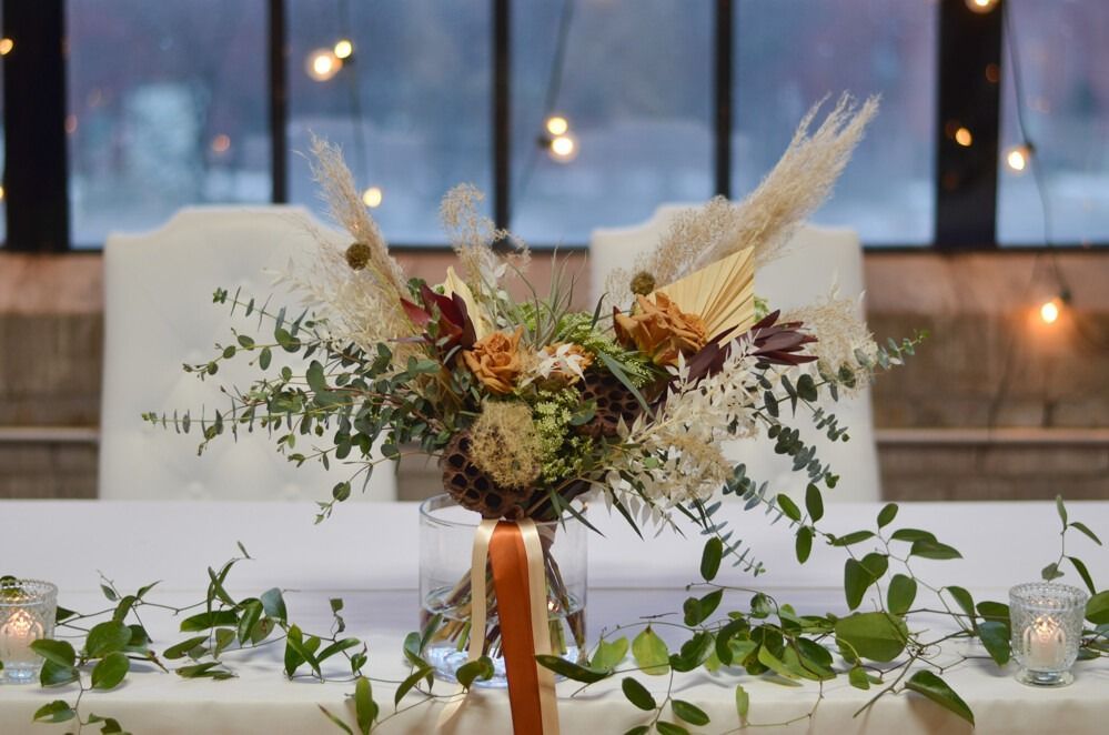 Floral centerpiece on table with greenery, candles, and background lights.