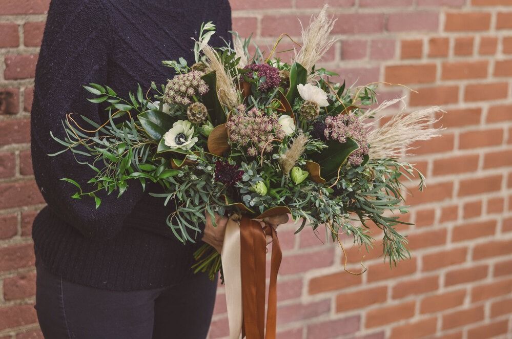 Person holding a bouquet of flowers in front of a brick wall. The flowers have green and white hues, and the bouquet is wrapped in brown paper and ribbon.