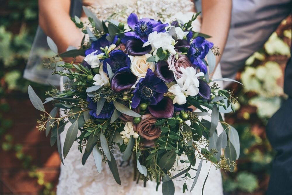 Bride holding a lush bouquet of blue, white, and purple flowers with greenery; outdoor setting.