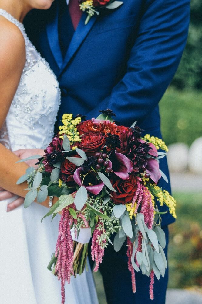 Wedding bouquet with burgundy, purple, and yellow flowers held by a bride and groom in a blue suit.