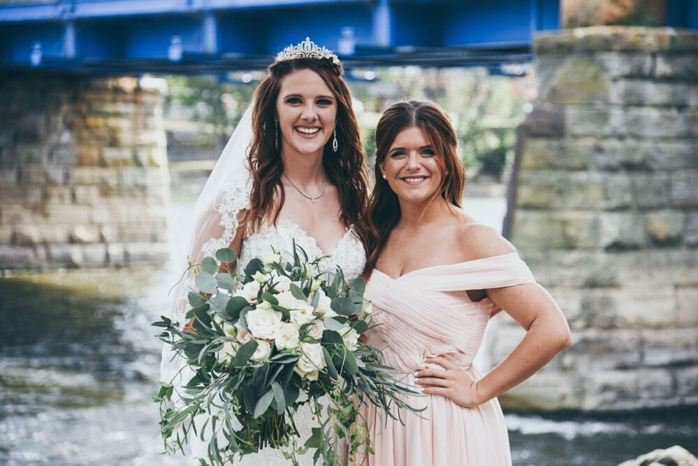 Bride with bouquet and bridesmaid in light pink dress smiling outdoors near a blue bridge.
