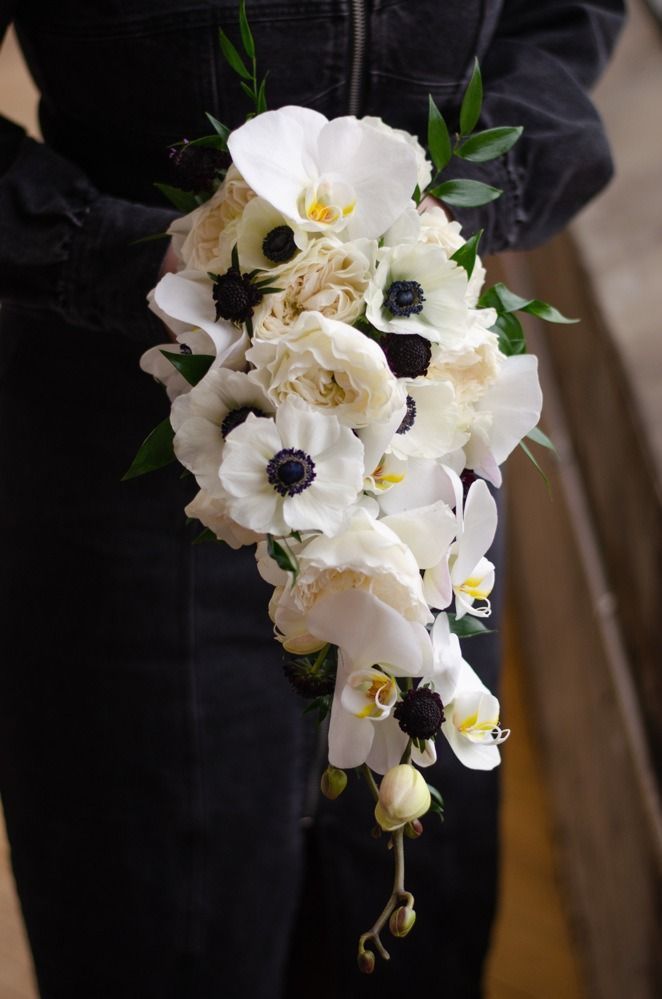 Person holding a cascading white floral bouquet with orchids and anemones against a dark background.