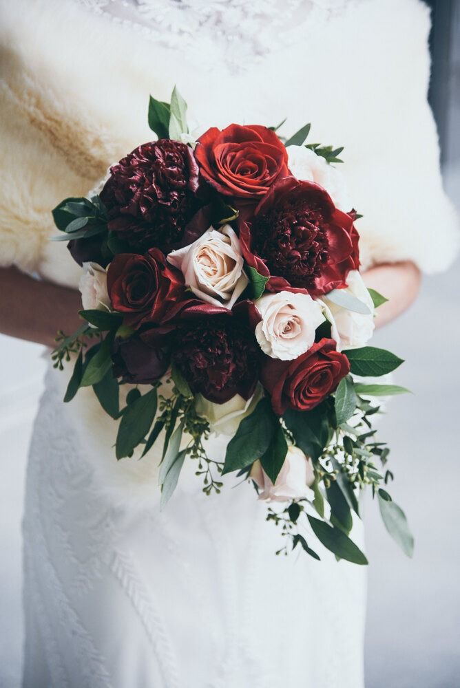 Bride holding bouquet of red and blush roses, wearing a fur stole and white dress.
