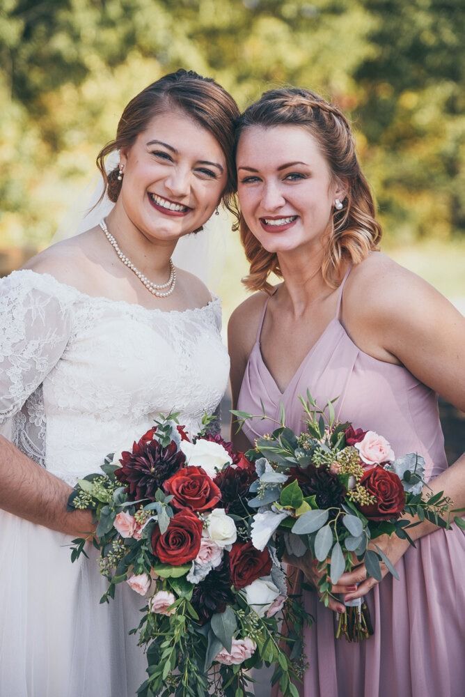 Bride and bridesmaid smiling, holding bouquets, standing outdoors. The bride wears a white lace dress; the bridesmaid wears a pink dress.