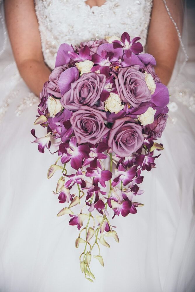 Bride holding a cascading purple rose and orchid bouquet, white wedding dress.