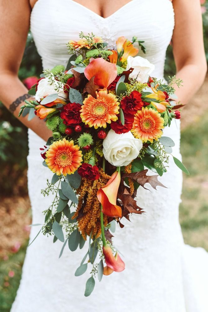 Bride in white dress holding autumn-colored cascading bouquet with orange, red, and white flowers.