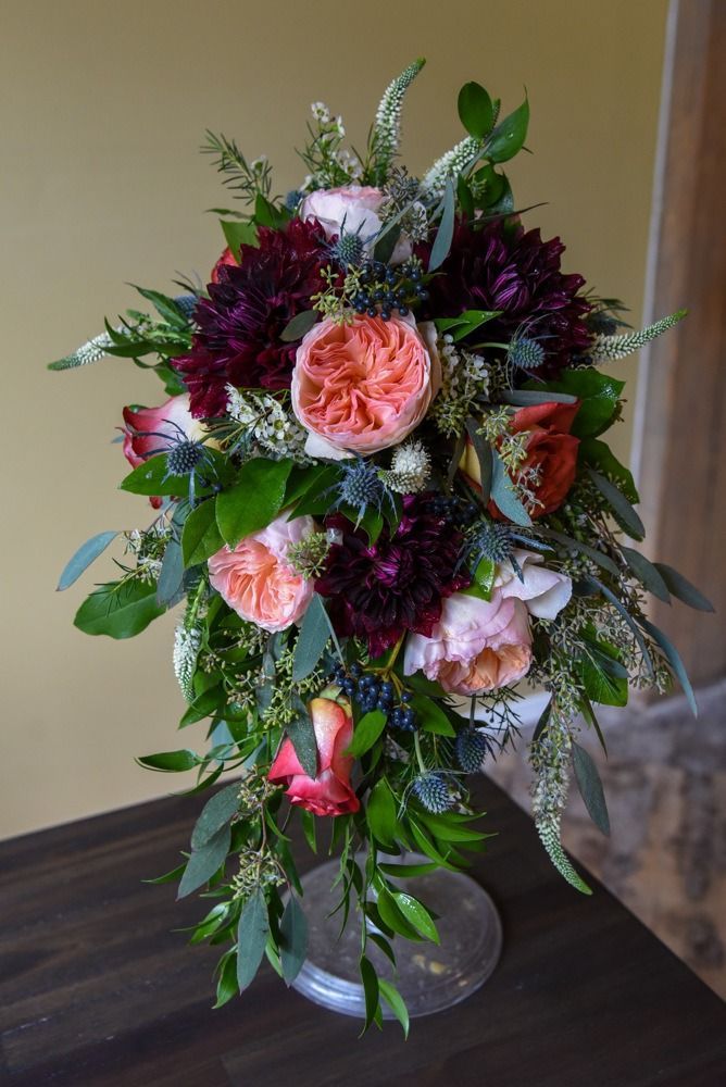 Cascading floral arrangement; coral and burgundy blooms, green foliage, on dark table, yellow backdrop.