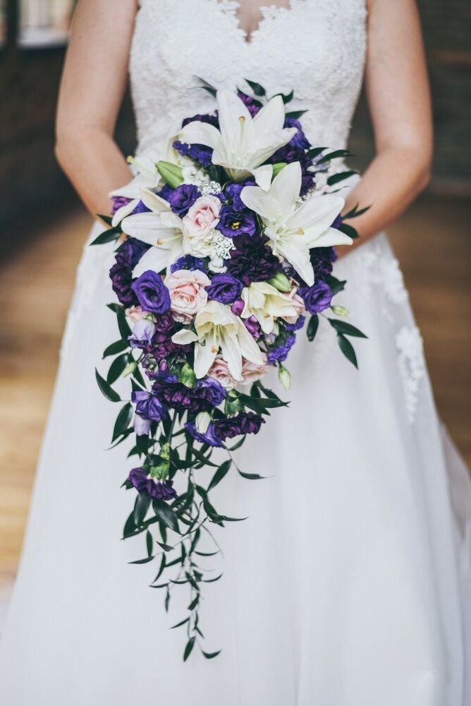 Bride in white dress holding cascading purple and white floral bouquet.