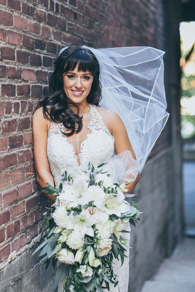 Bride in lace dress leans against a brick wall, holding a bouquet, veil blowing.