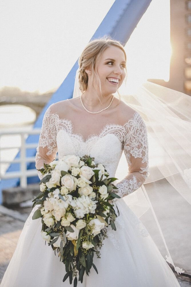 Bride in white lace dress smiles, holding bouquet. Long veil blows in the wind, standing on a bridge.