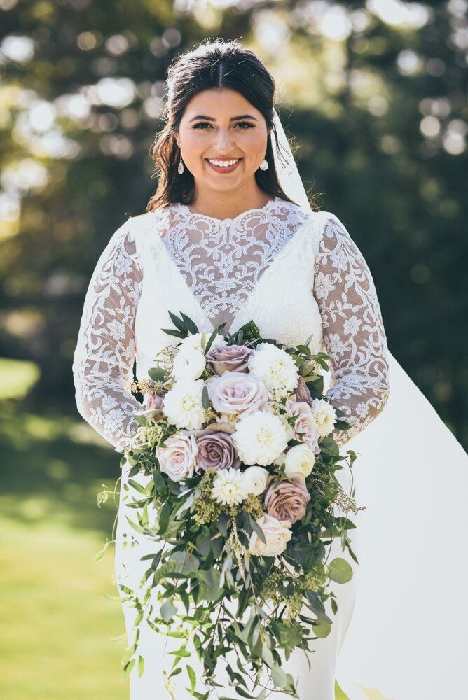 Bride in white lace wedding dress, smiling, holding bouquet, outdoor setting.