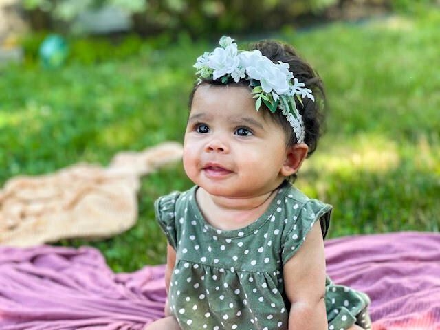 Baby wearing a white floral headband, smiles while sitting on a purple blanket in a green yard.
