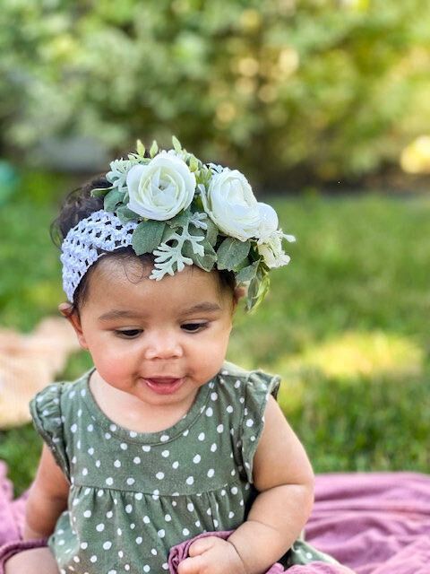 Baby with a flower crown and green polka-dot dress smiles outdoors, seated on a pink blanket, near foliage.