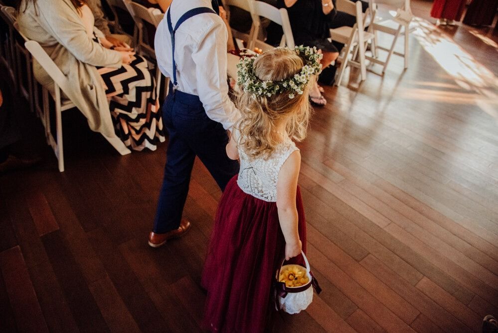 Bridesmaid in a burgundy dress with a flower crown walks down the aisle, holding hands with a man wearing suspenders.