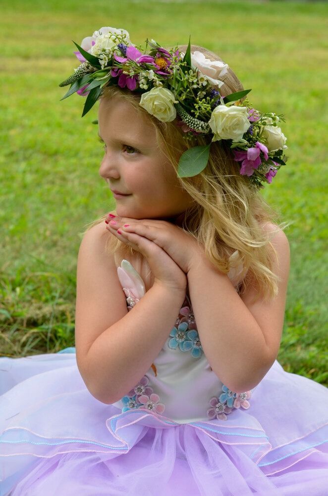 Girl in floral crown and pastel dress, hands clasped, looking thoughtful on grass.