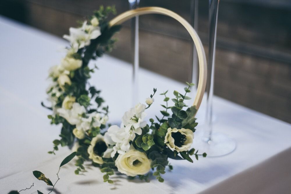 Floral wreath on a gold ring, with white and green flowers, next to glass vases on a white surface.