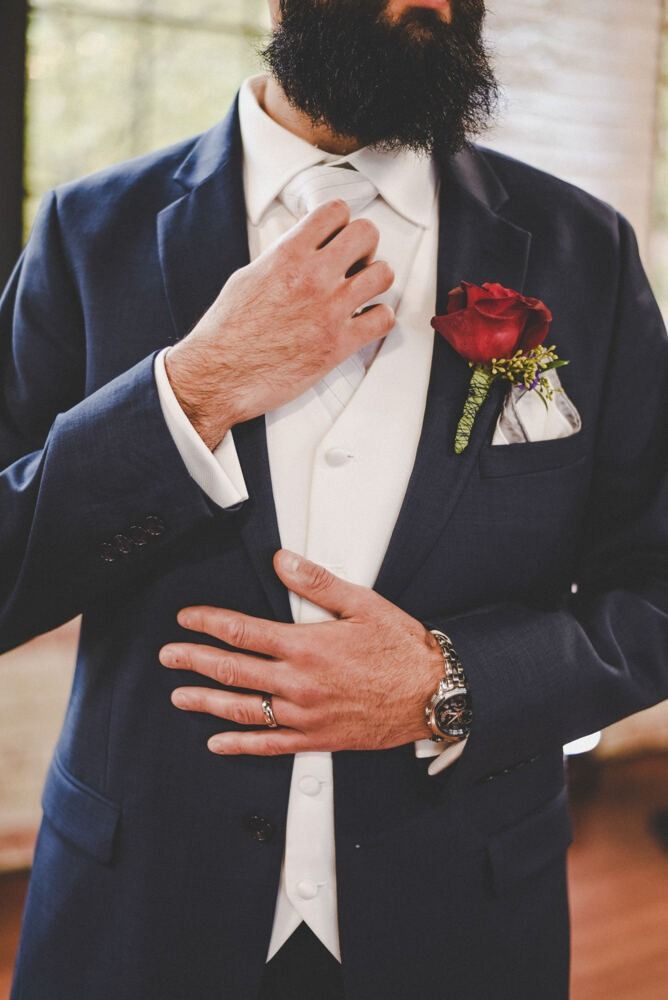 Man in navy suit adjusting tie, wearing a white vest, with a red rose boutonniere.