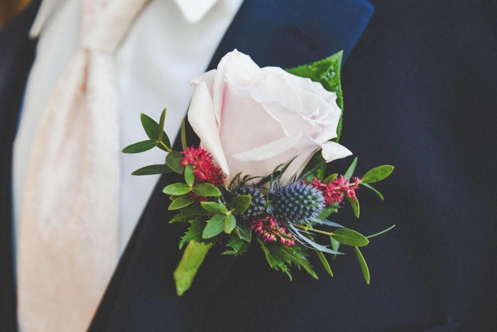 Boutonniere of a light pink rose, blue thistle, and red accents on a navy suit.