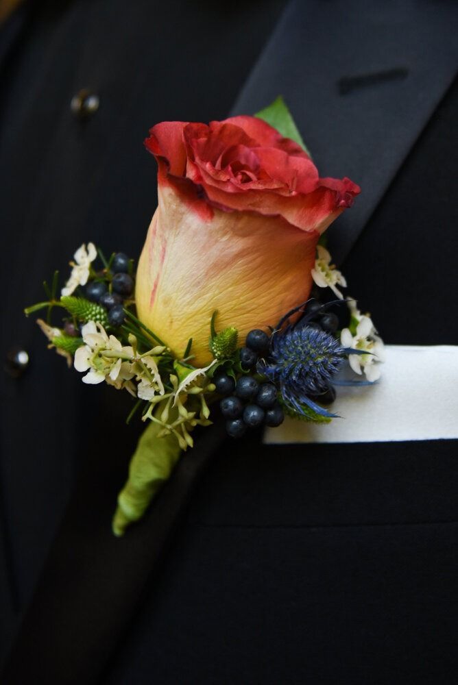 Boutonniere with red-yellow rose, blue berries, and thistle on black suit.