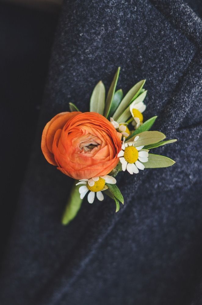 Orange flower boutonniere with white daisies and green leaves on a dark gray suit jacket.