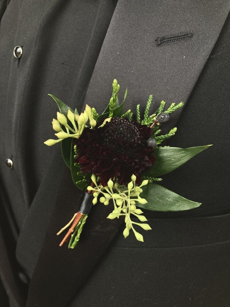 Black boutonniere with dark burgundy flower, greenery, and small yellow buds pinned to a black suit.