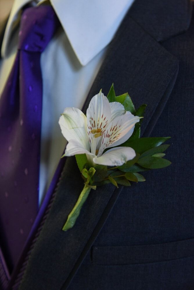 White flower boutonniere with green leaves on a dark suit, purple tie visible.
