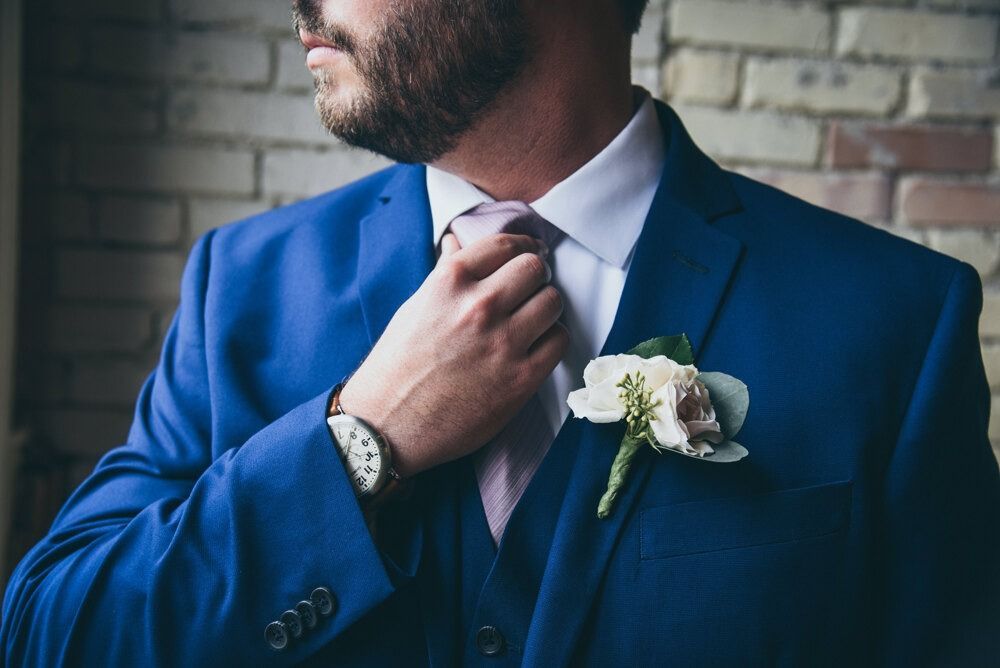 Man in blue suit adjusting tie, wearing watch, flower boutonniere on jacket, against brick wall.
