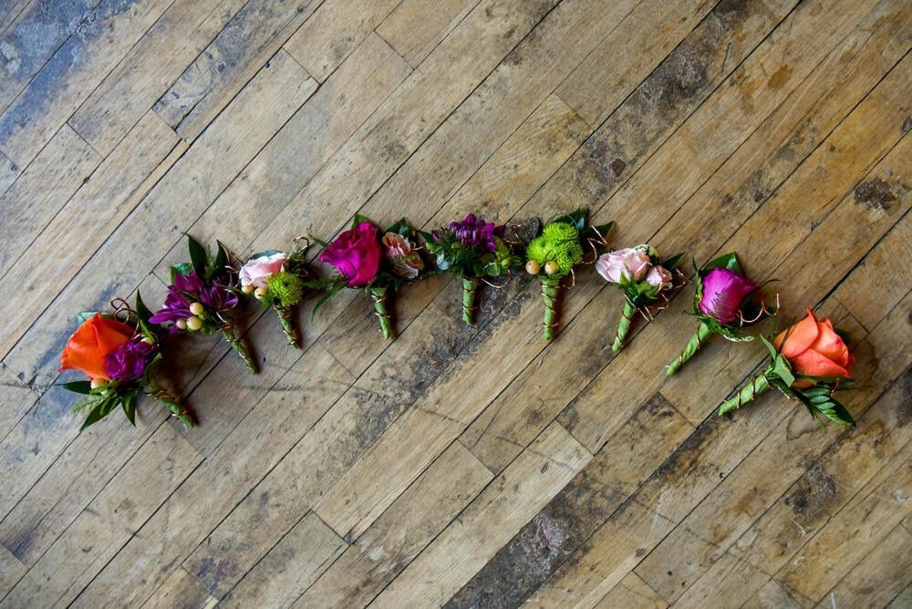 Row of colorful boutonnieres on a wooden floor, featuring roses and greenery.