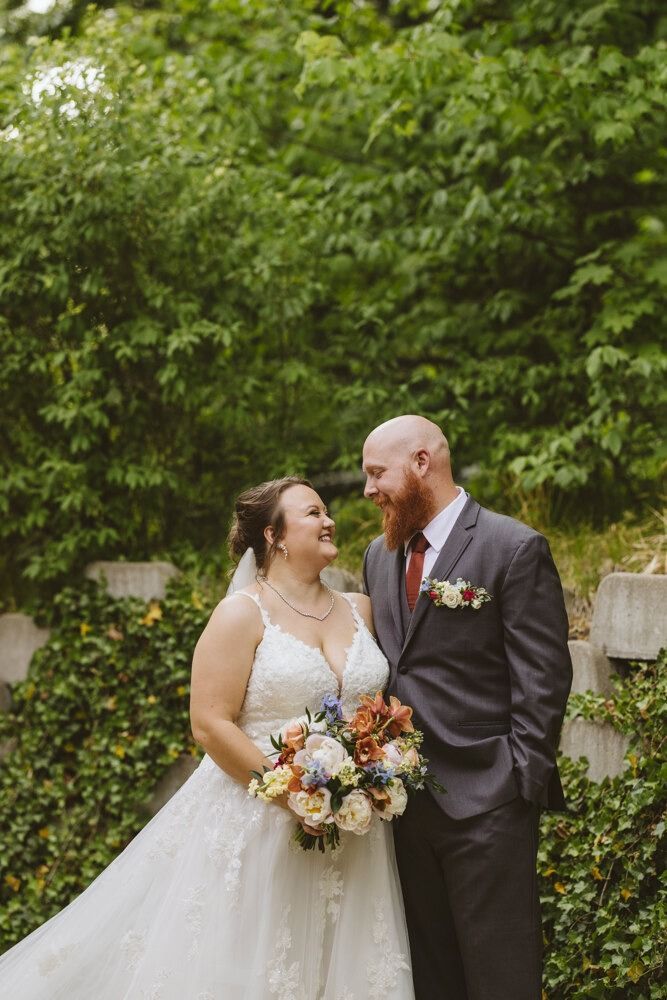 Bride and groom smiling at each other, holding flowers, in front of a green wall of foliage.