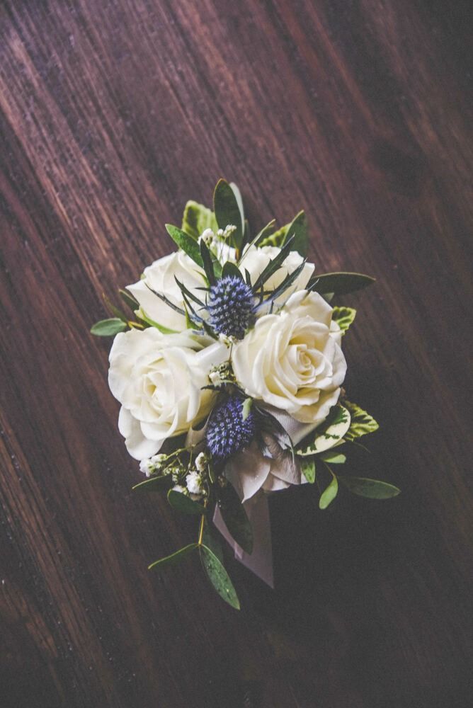 White rose and blue thistle boutonniere on a dark wooden surface, with greenery.