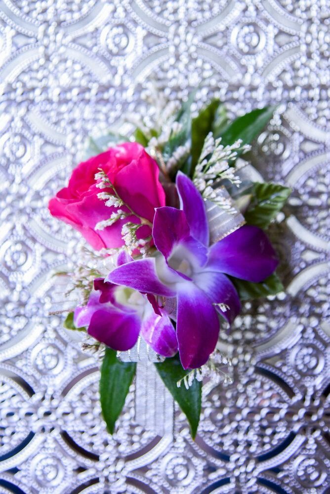 Close-up of a corsage with a pink rose and purple orchids on a patterned silver background.