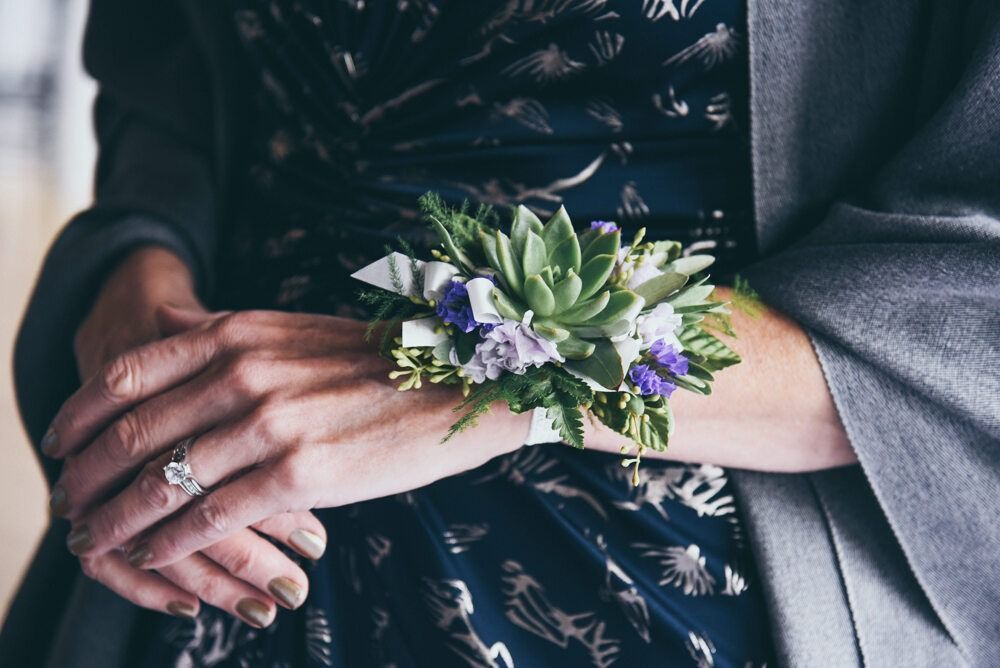 Woman's hands holding a floral wrist corsage with succulents and purple accents, wearing a dark dress and grey shawl.