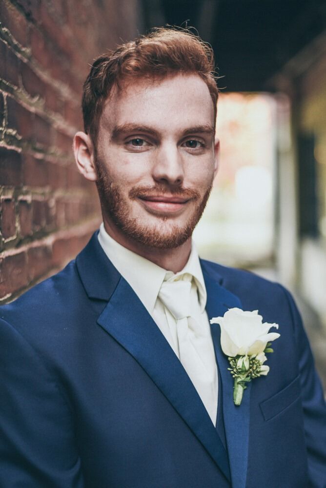 Man in navy suit, white tie and rose, smiling in front of a brick wall.