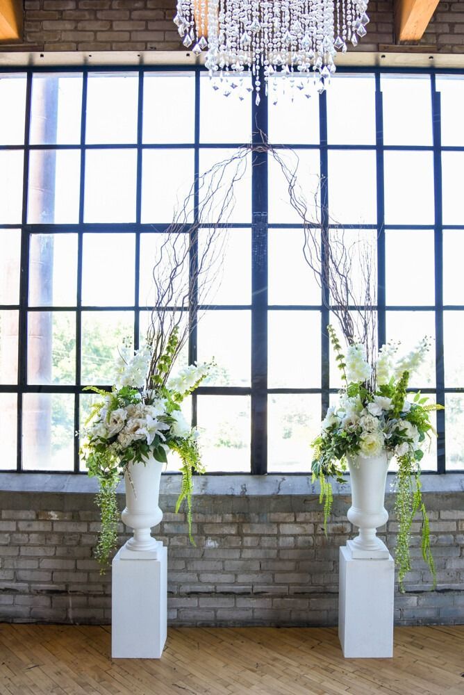 Wedding ceremony backdrop: white urn floral arrangements, branches, and large window in a brick building.