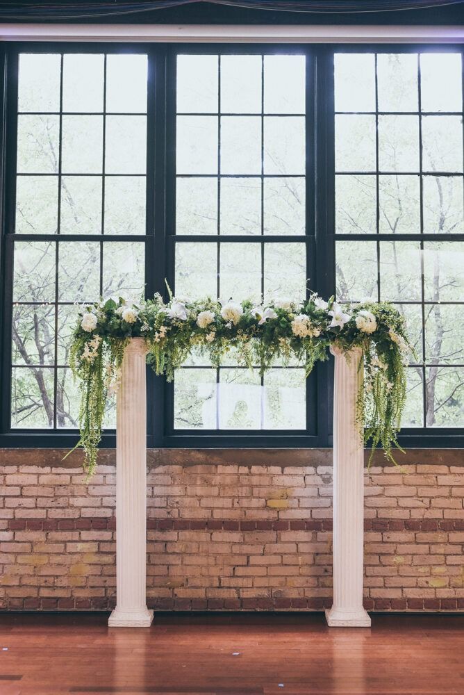 Floral arch on white columns in front of a window and brick wall, setting for an event.
