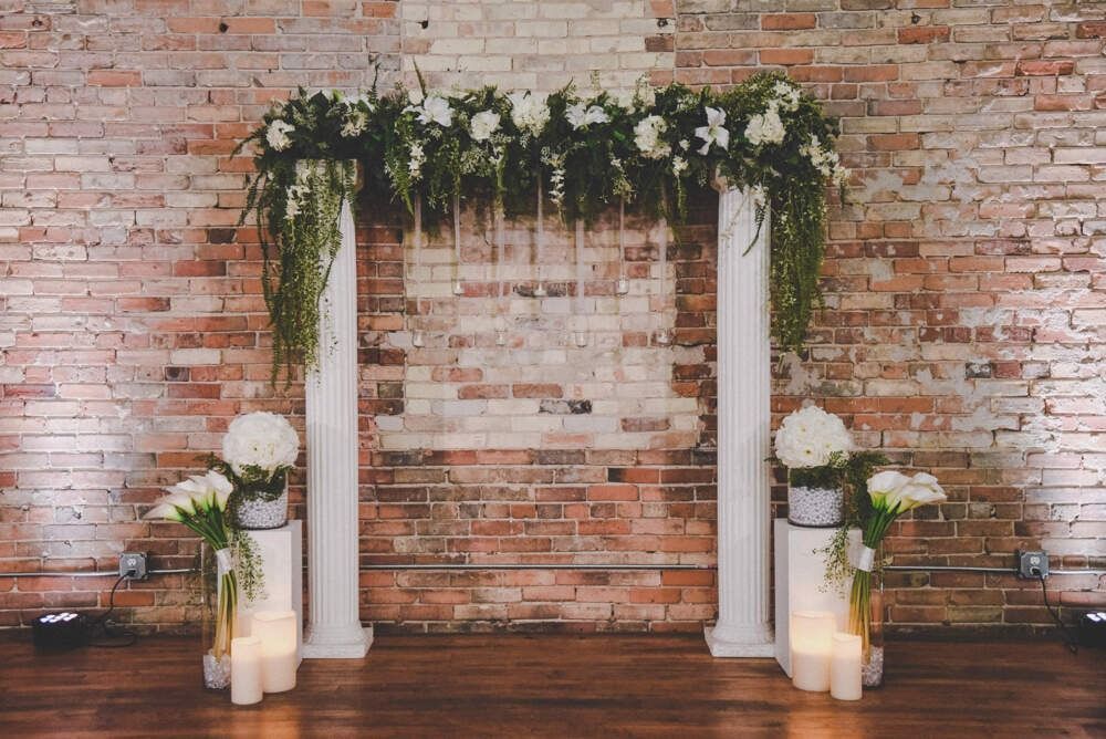 Floral wedding archway with white pillars against a brick wall.