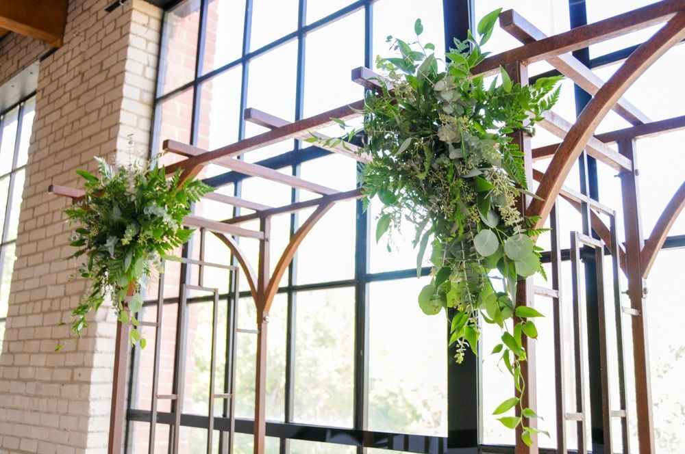 Wedding arch with green foliage and white flowers against large windows.
