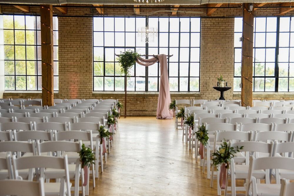 Wedding ceremony setup in a brick building with rows of white chairs facing an arch decorated with flowers and fabric.