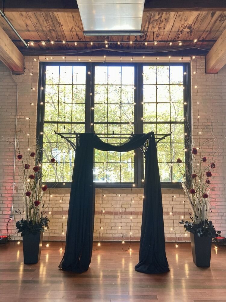 Wedding arch with draped black fabric and string lights in front of a window.