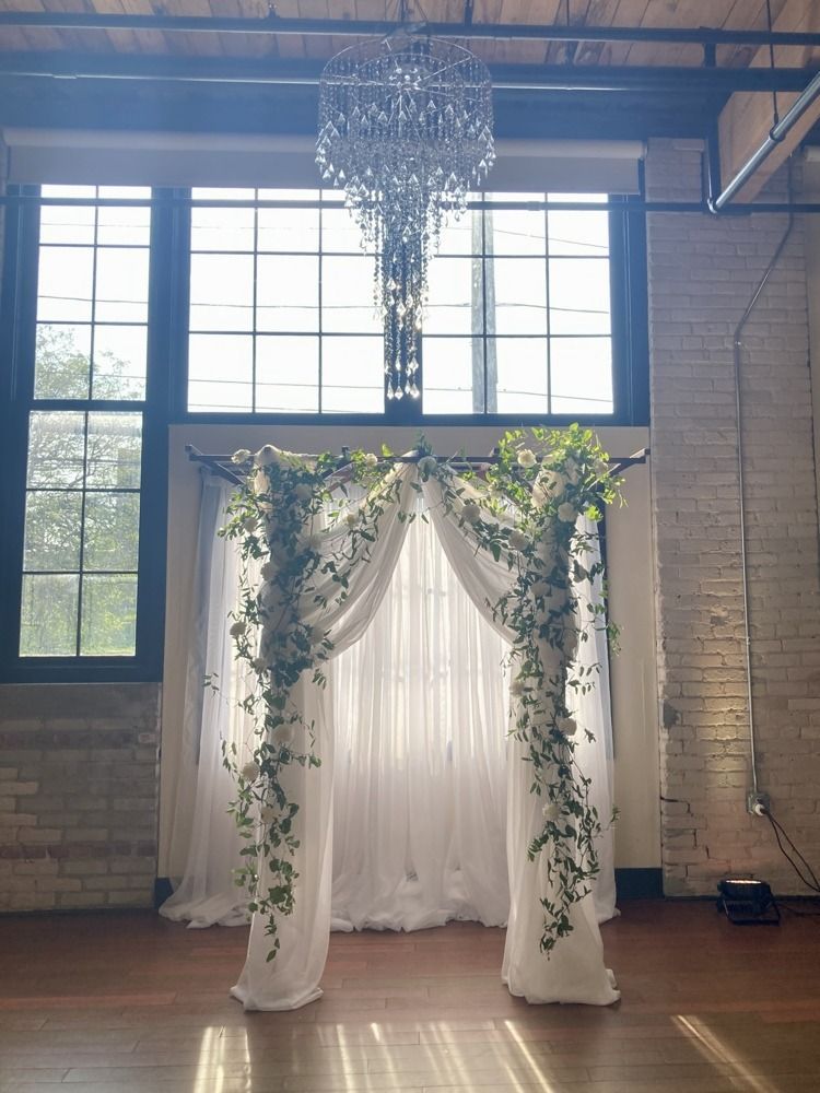 Wedding arch draped with white fabric and flowers, in front of a window and under a chandelier.
