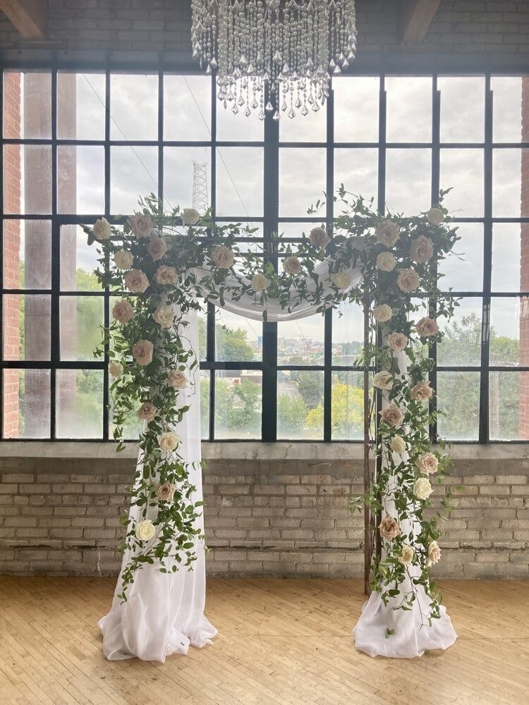 Wedding arch decorated with flowers and greenery in front of a large window, under a chandelier.
