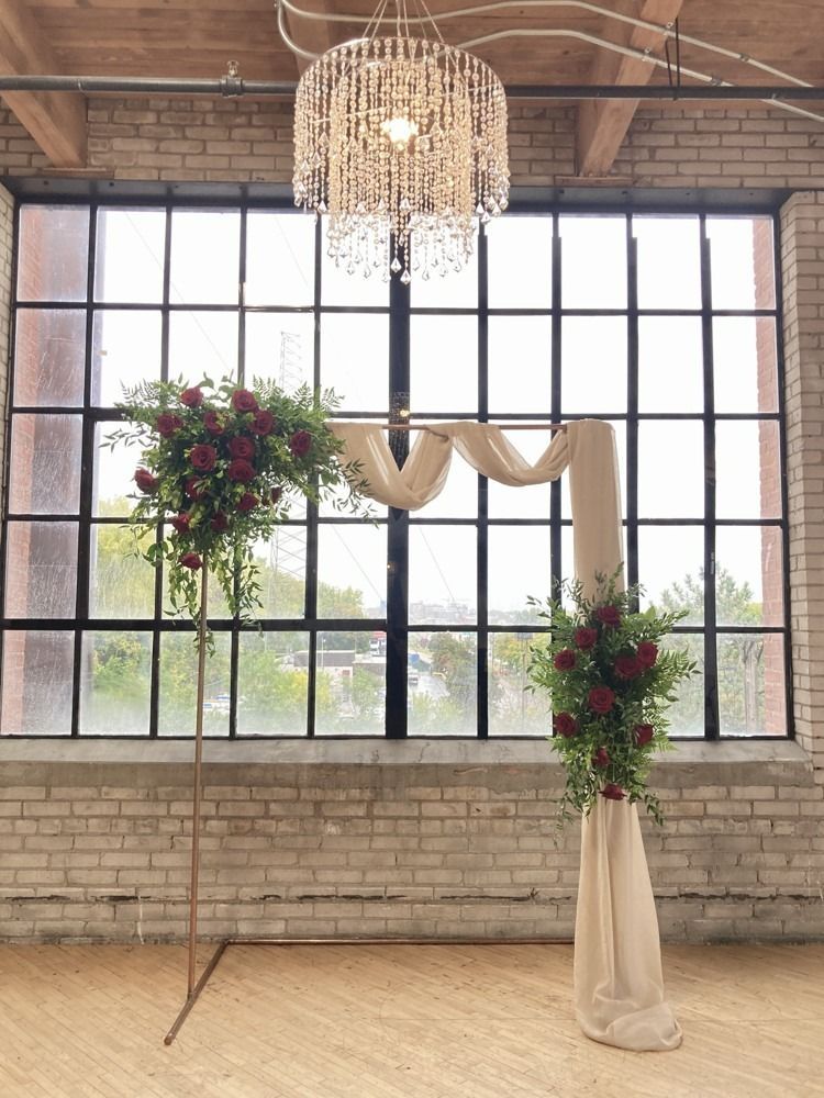 Wedding arch with burgundy flowers, draped fabric, and a chandelier in front of a window.