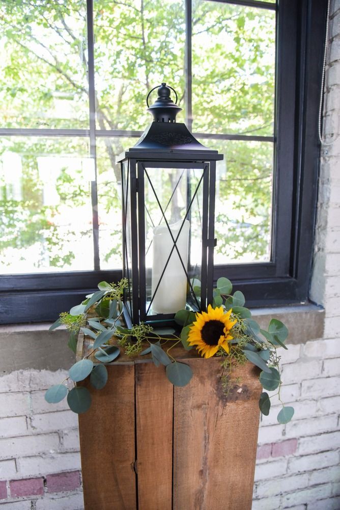 Black lantern with lit candle on a wooden stand, decorated with greenery and a sunflower, in front of a window.