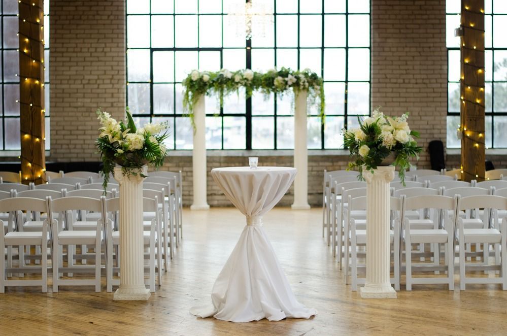 Wedding ceremony setup in a loft with white chairs, columns, and floral arch.